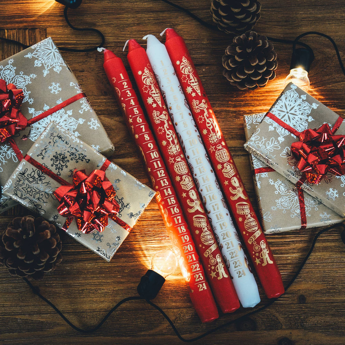 Four Christmas countdown candles in red and white with gold festive illustrations, arranged on a wooden table alongside wrapped gifts, pinecones, and warm string lights.