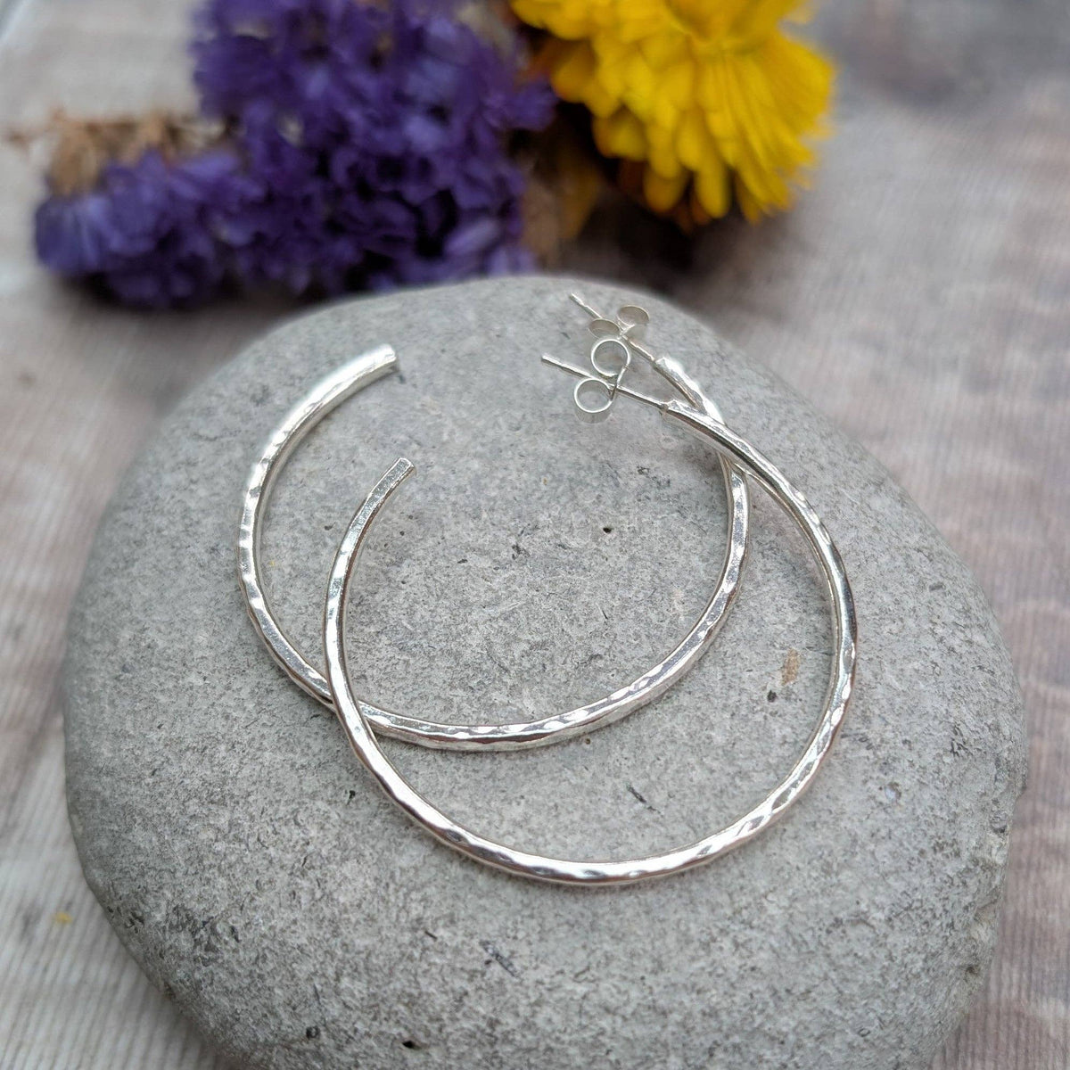 Silver hoop earrings with a hammered texture, displayed on a round gray stone with dried purple and yellow flowers in the background.