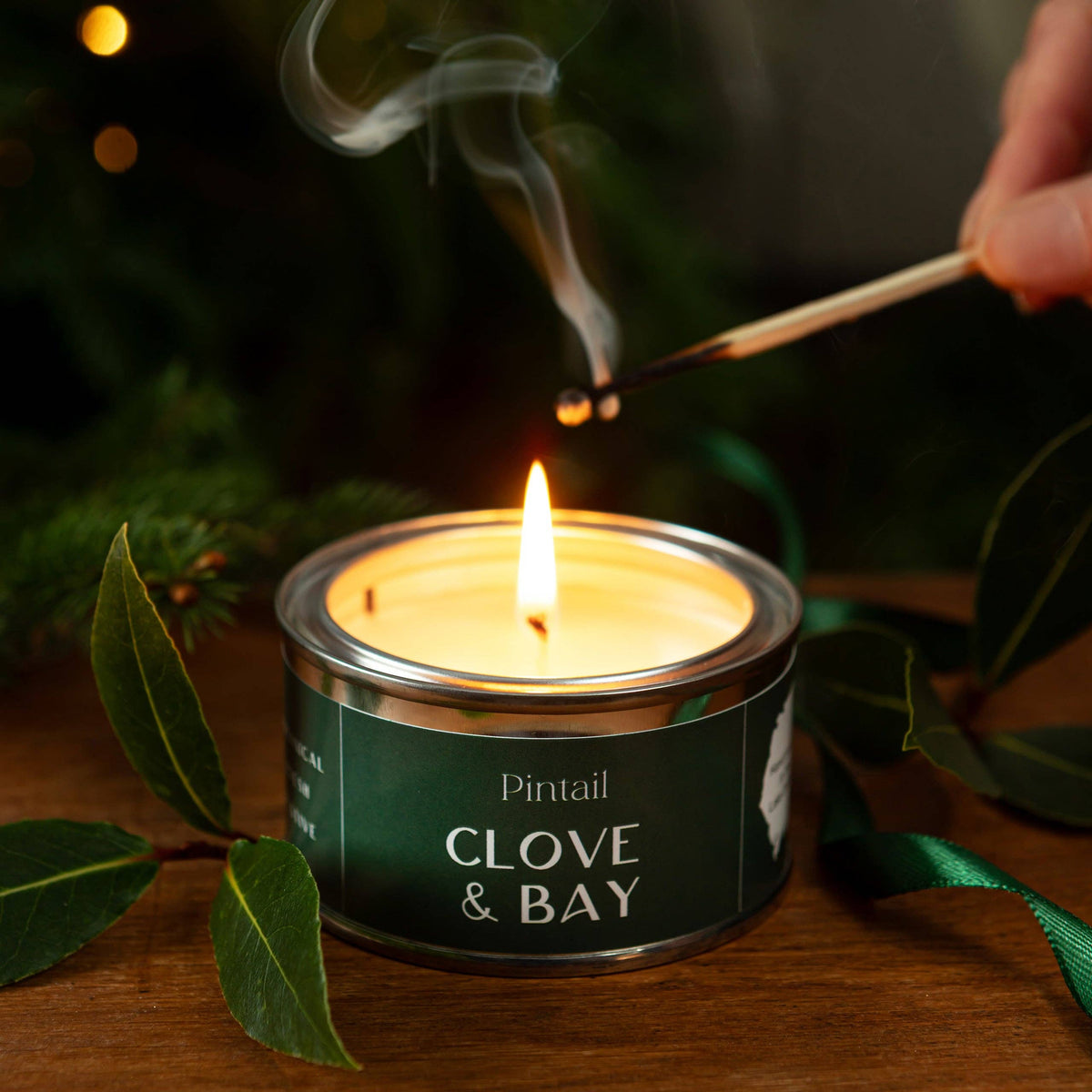 Candle labeled 'Clove & Bay' being lit with a match, surrounded by greenery on a wooden surface.
