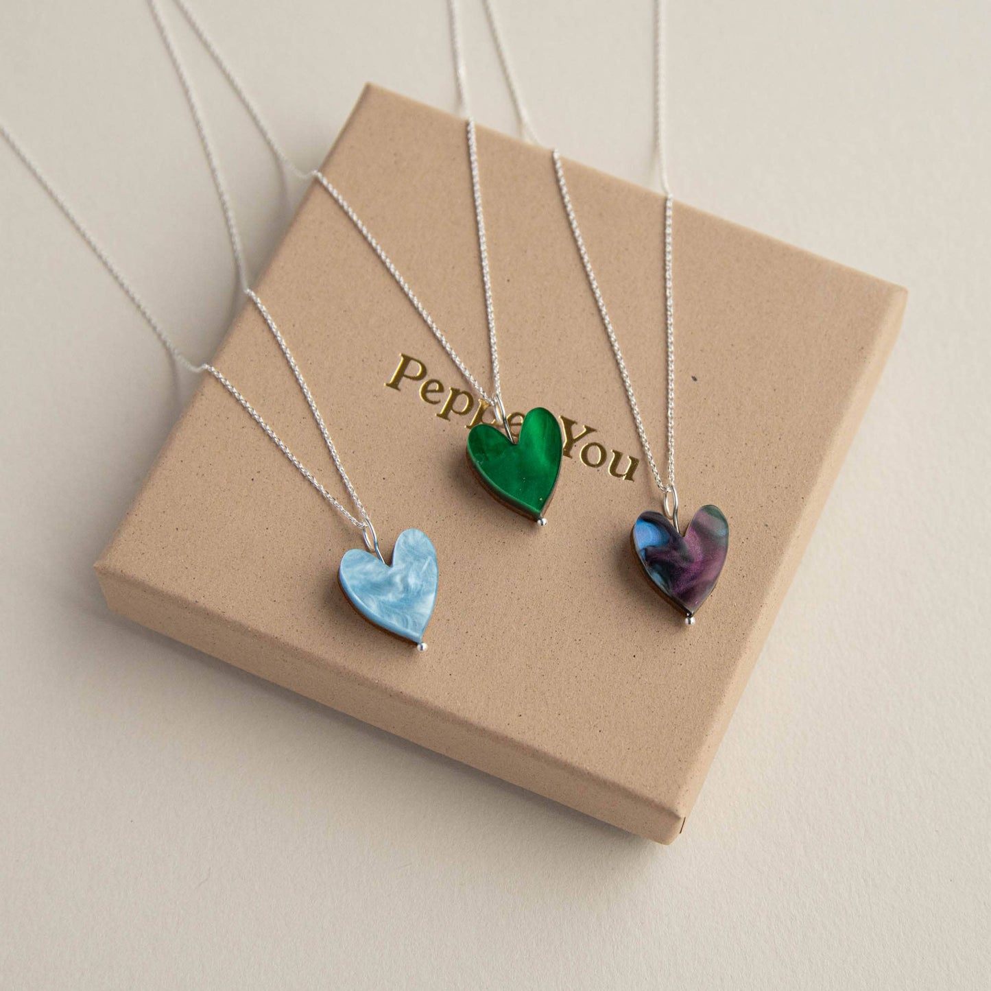 Three heart-shaped pendants on necklaces displayed on a 'Pepper You' box.