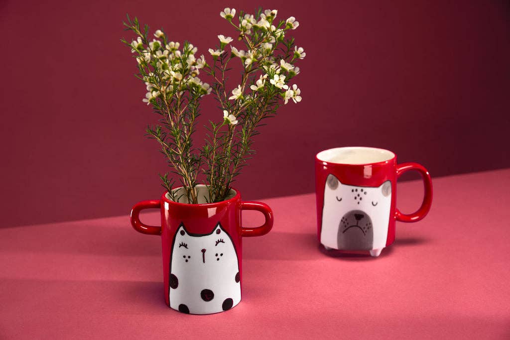Two red mugs with cat designs on a pink surface against a red background
