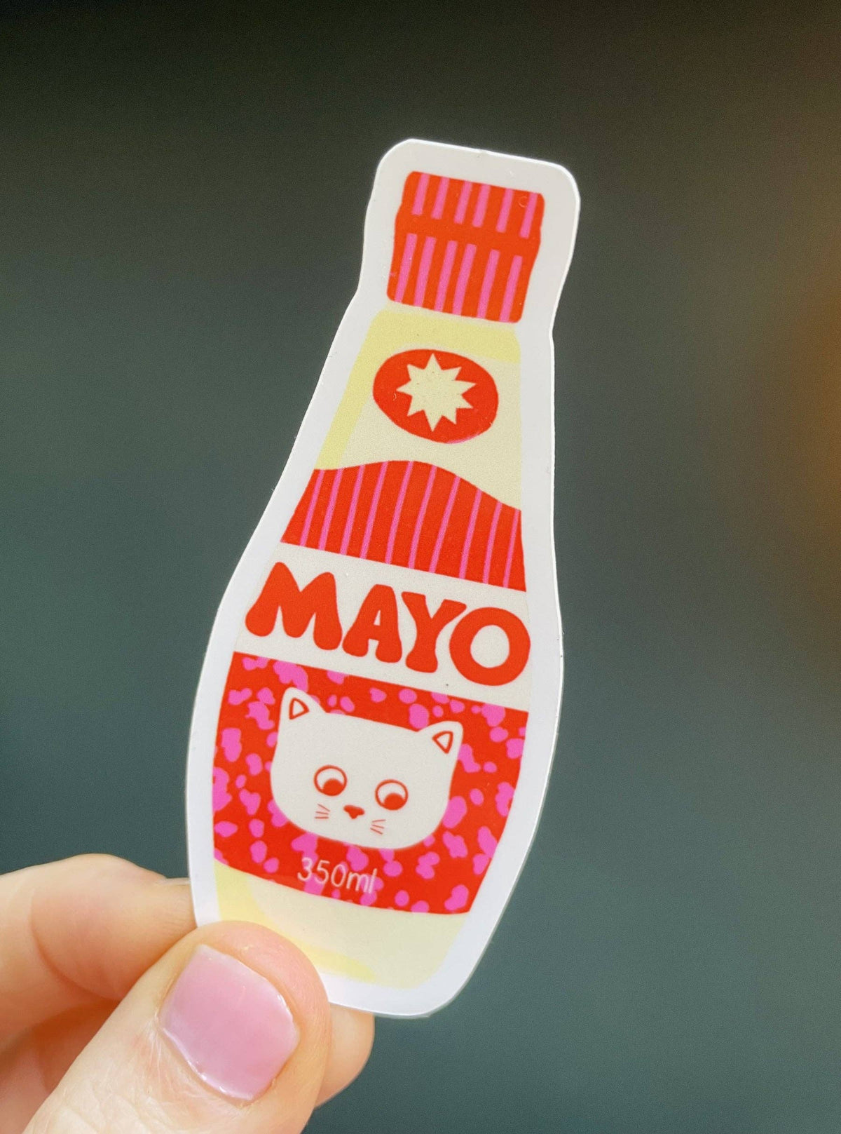 Sticker of a mayo bottle with a cat face design held by a hand against a dark background