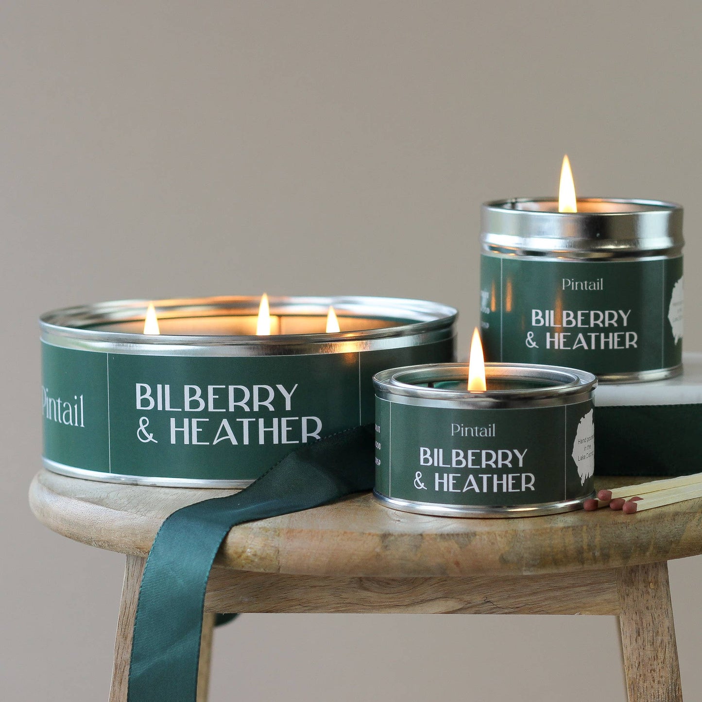 Three candles in green tins labeled 'Bilberry & Heather' on a wooden stand.