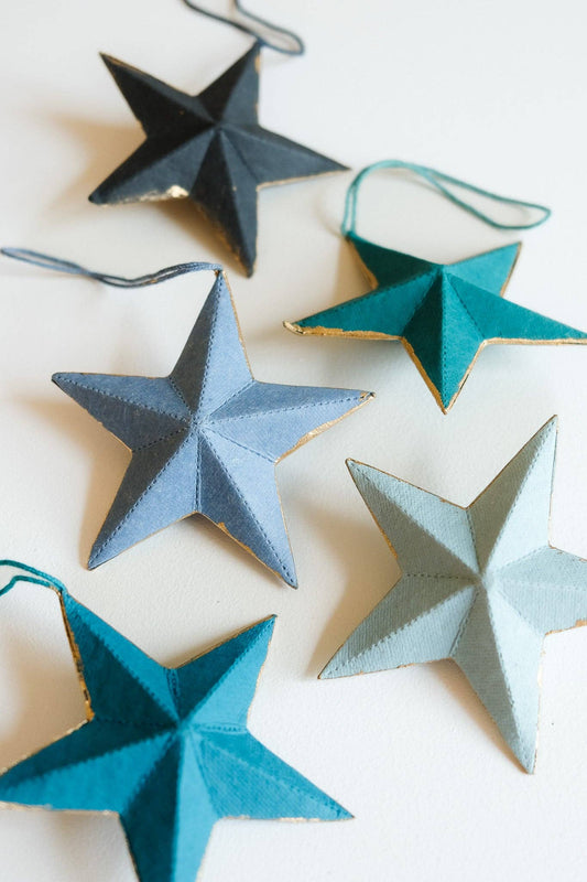 Five handmade paper star ornaments in various shades of blue, teal, and light gray, arranged on a plain white background.