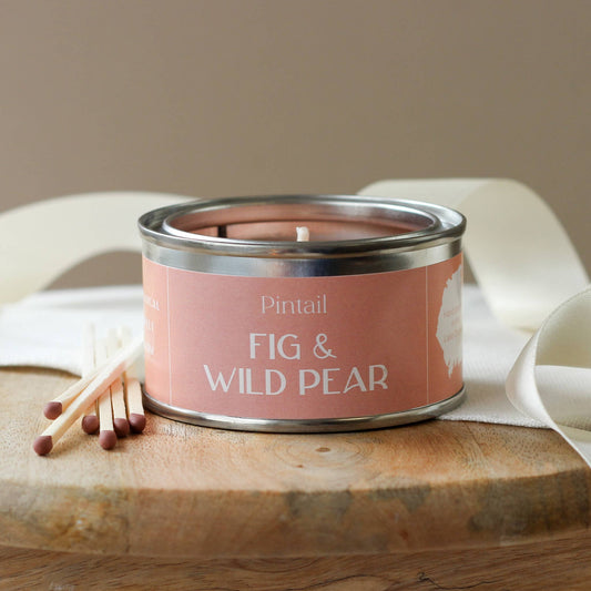 A Fig and Wild Pear scented candle in a metal paint pot tin with a lit flame, placed on a wooden surface with white linen and match sticks beside it.