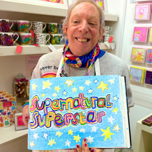 Person holding a book titled 'Supernatural Superstar' in a store setting with colorful items on shelves.