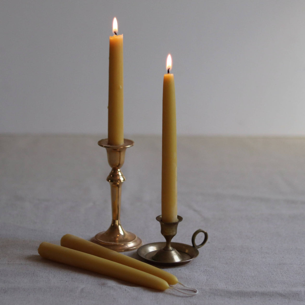 Two lit candles in brass candlesticks with two unlit candles on a plain background