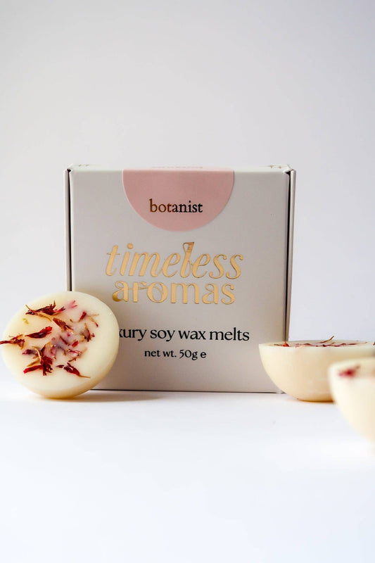A box of Botanist Timeless Aromas luxury soy wax melts with two round wax melts shown in the foreground, one cut in half to reveal dried botanical pieces inside, on a clean white background.
