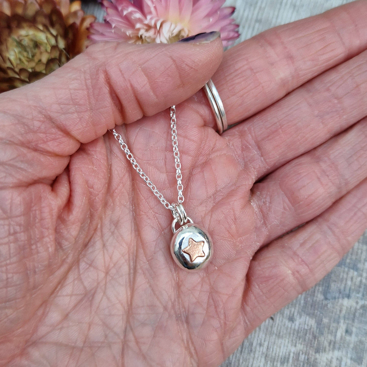 A silver necklace with a small round pendant featuring a star design, held in a person's hand. The person is also wearing a silver ring.