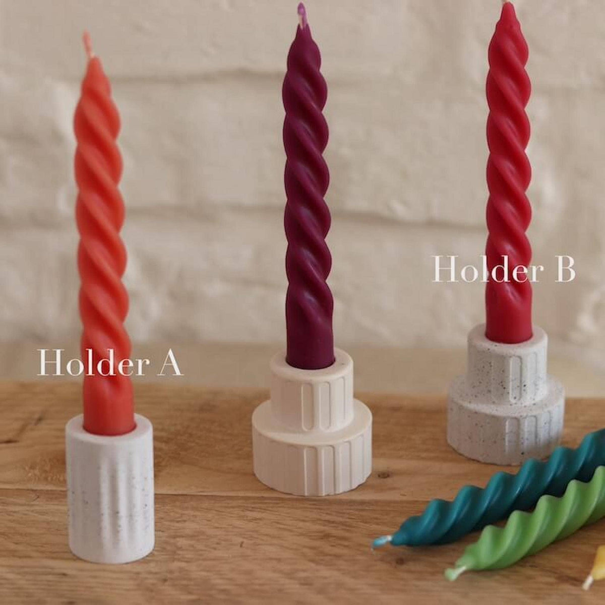 Three twisted spiral taper candles in red, dark purple, and red, each mounted in a white plastic holder on a wooden surface against a light brick wall. Two additional teal and green spiral candles are lying on the right, with text labels 'Holder A', 'Holder B', and 'Holder C' overlaid.