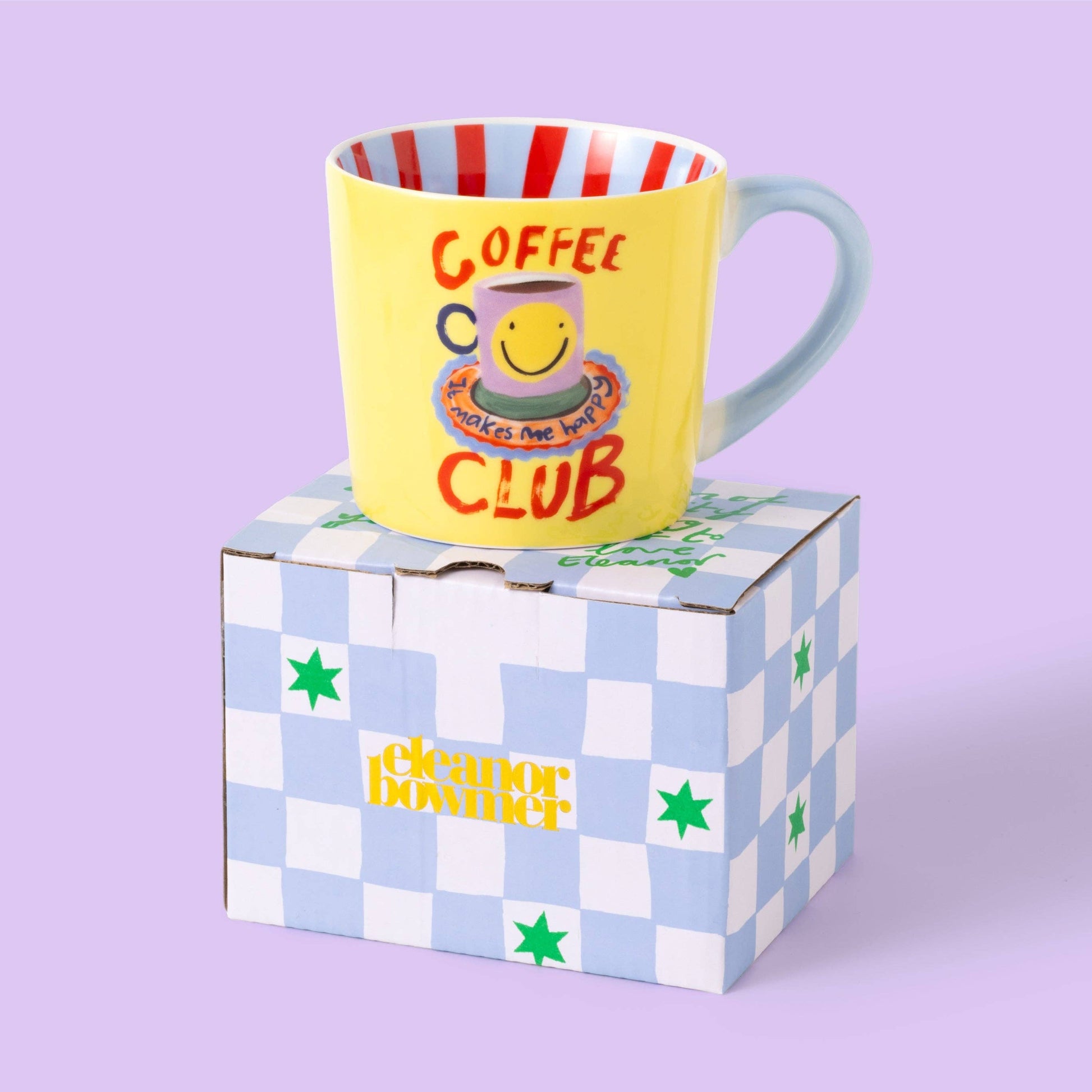 Yellow mug with red text and smiley face design on a checkered box against a purple background
