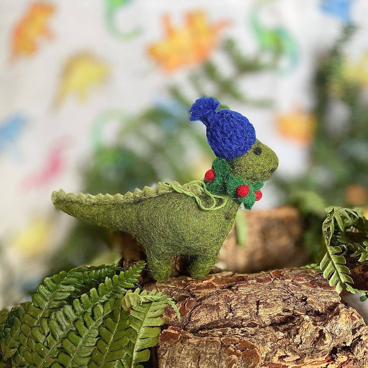 A small felt dinosaur figurine in olive green, wearing a blue knit hat and a green holly garland with red berries, posed on a piece of bark with ferns in the foreground.
