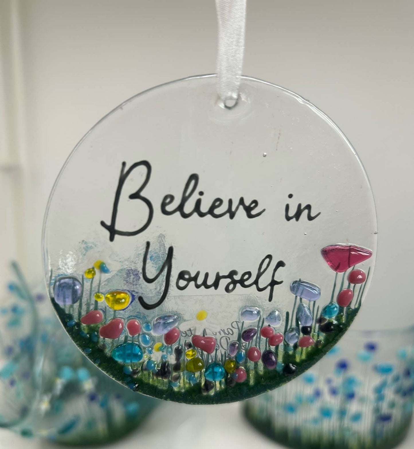 Round glass ornament with colorful flowers and 'Believe in Yourself' text on a white background