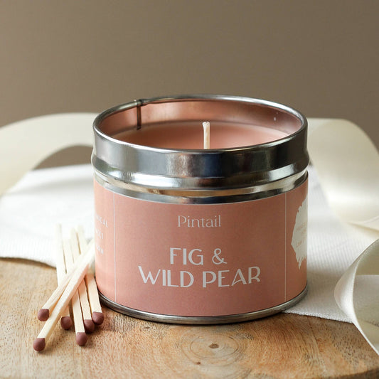 Candle in a pink tin labeled 'Fig & Wild Pear' by Pintail on a wooden surface with matches.