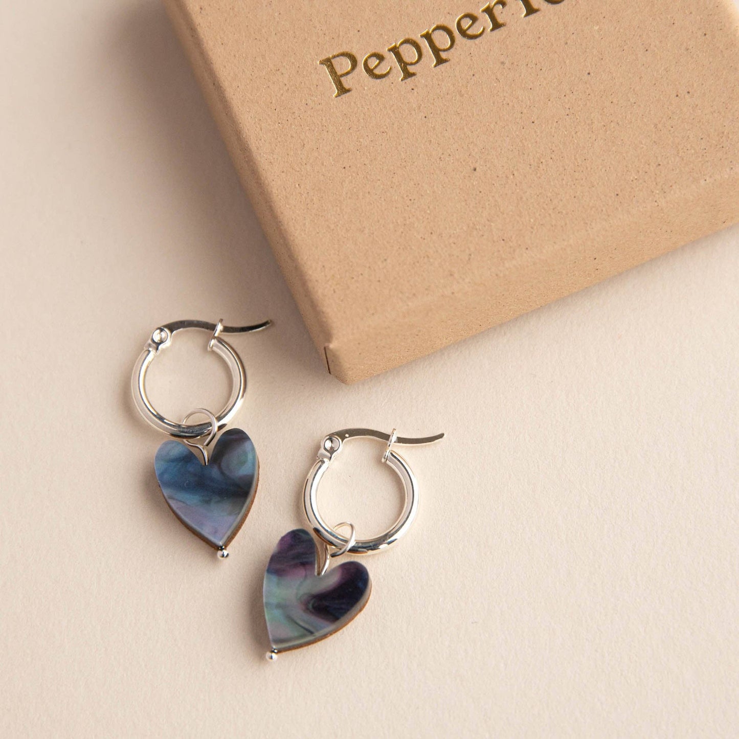Heart-shaped earrings with a metallic finish on a beige surface with 'Pepper' branding.