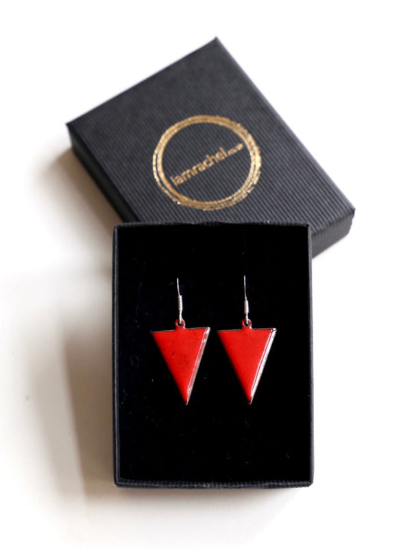 A pair of red inverse triangle earrings displayed in an open black box with a golden decorative border. The earrings are suspended from small silver hooks.