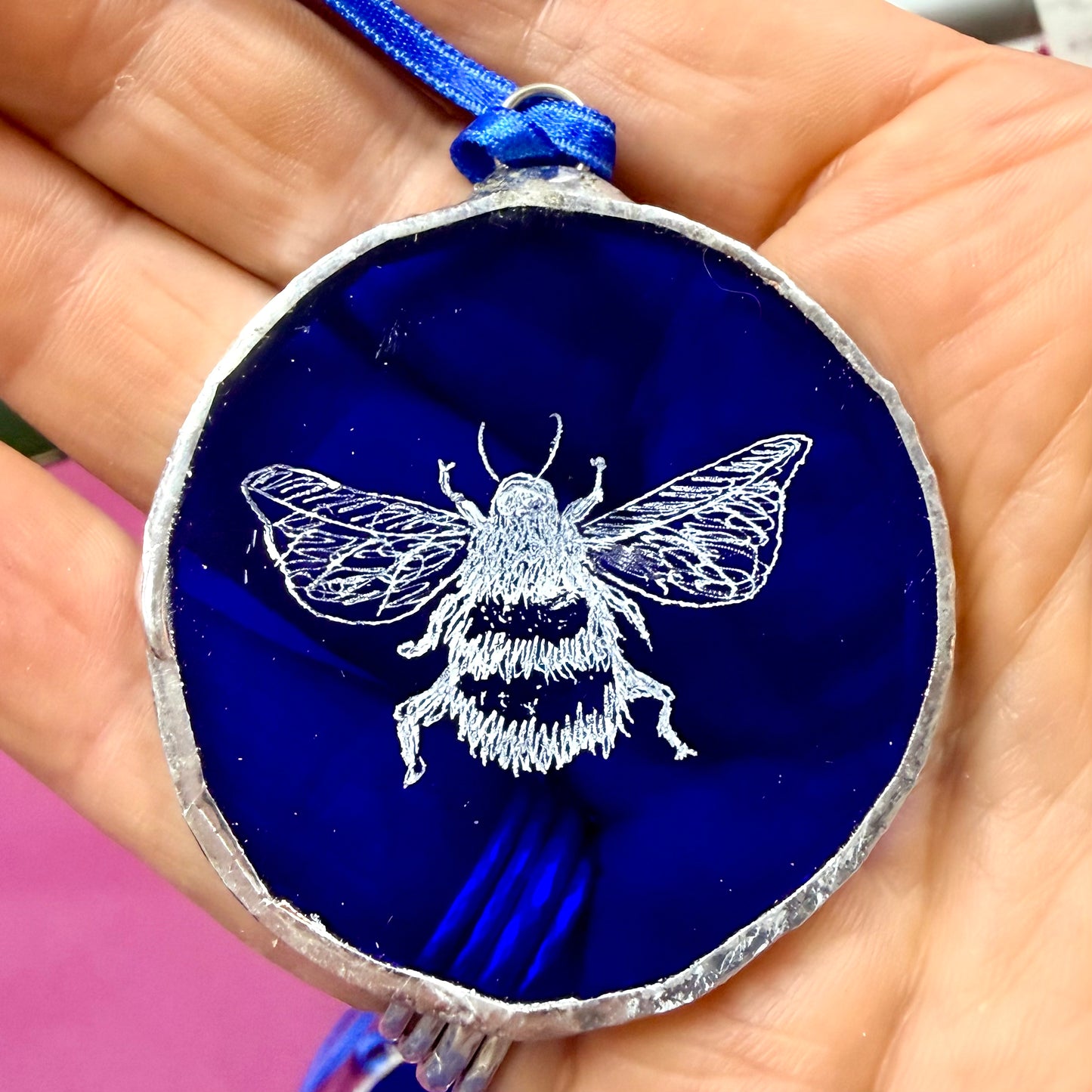 Blue glass disc with hoop and ribbon on a hand with a bumble bee etched into the glass