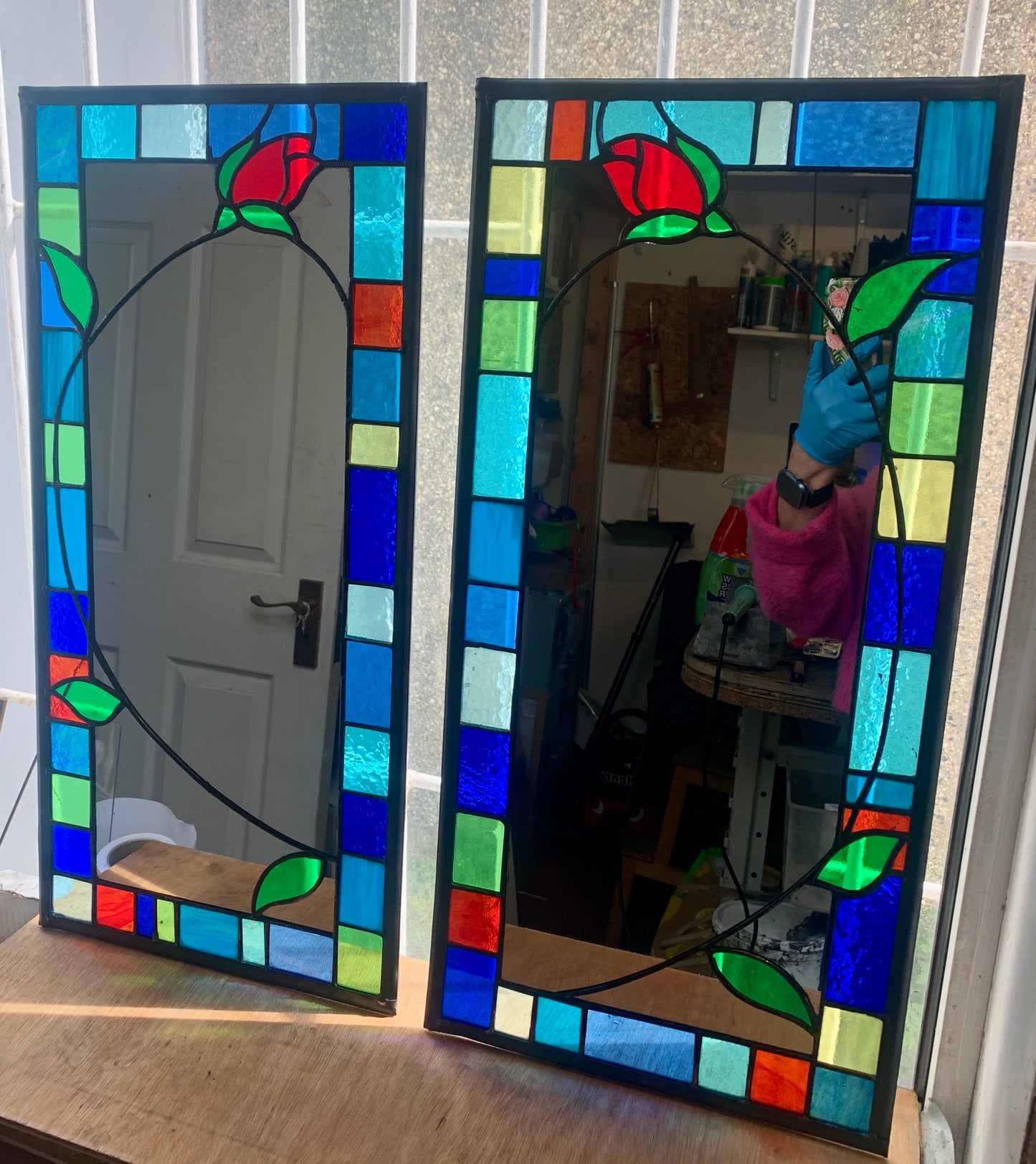 Tall slim stained glass mirror with random colours in various sizes in the border. Red rose bud detail with a long stem cut into the mirror glass and border. Copperwire attached to the back for hanging.