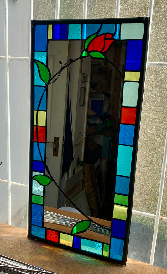 Tall slim stained glass mirror with random colours in various sizes in the border. Red rose bud detail with a long stem cut into the mirror glass and border. Copperwire attached to the back for hanging.