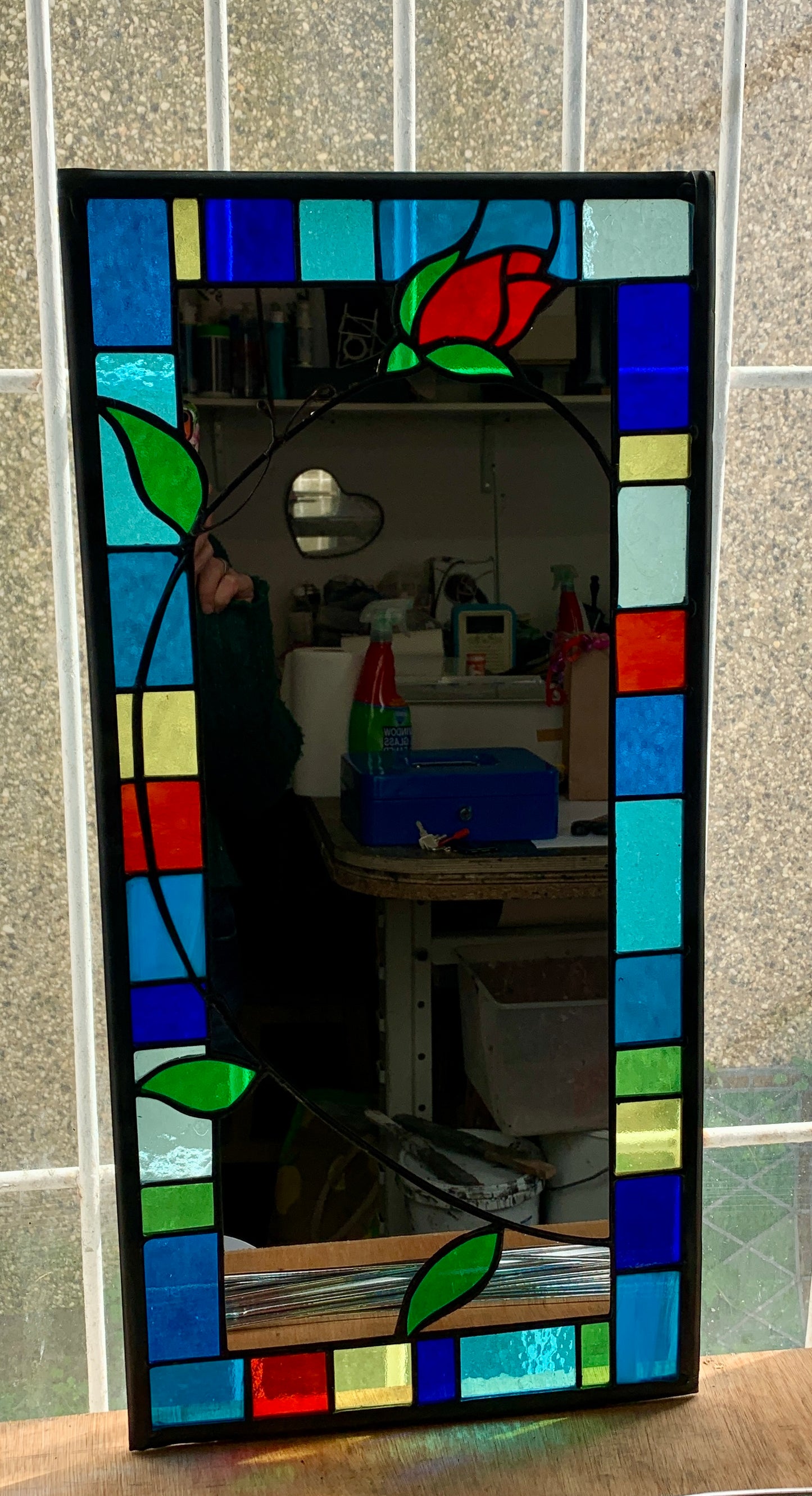 Tall slim stained glass mirror with random colours in various sizes in the border. Red rose bud detail with a long stem cut into the mirror glass and border. Copperwire attached to the back for hanging.