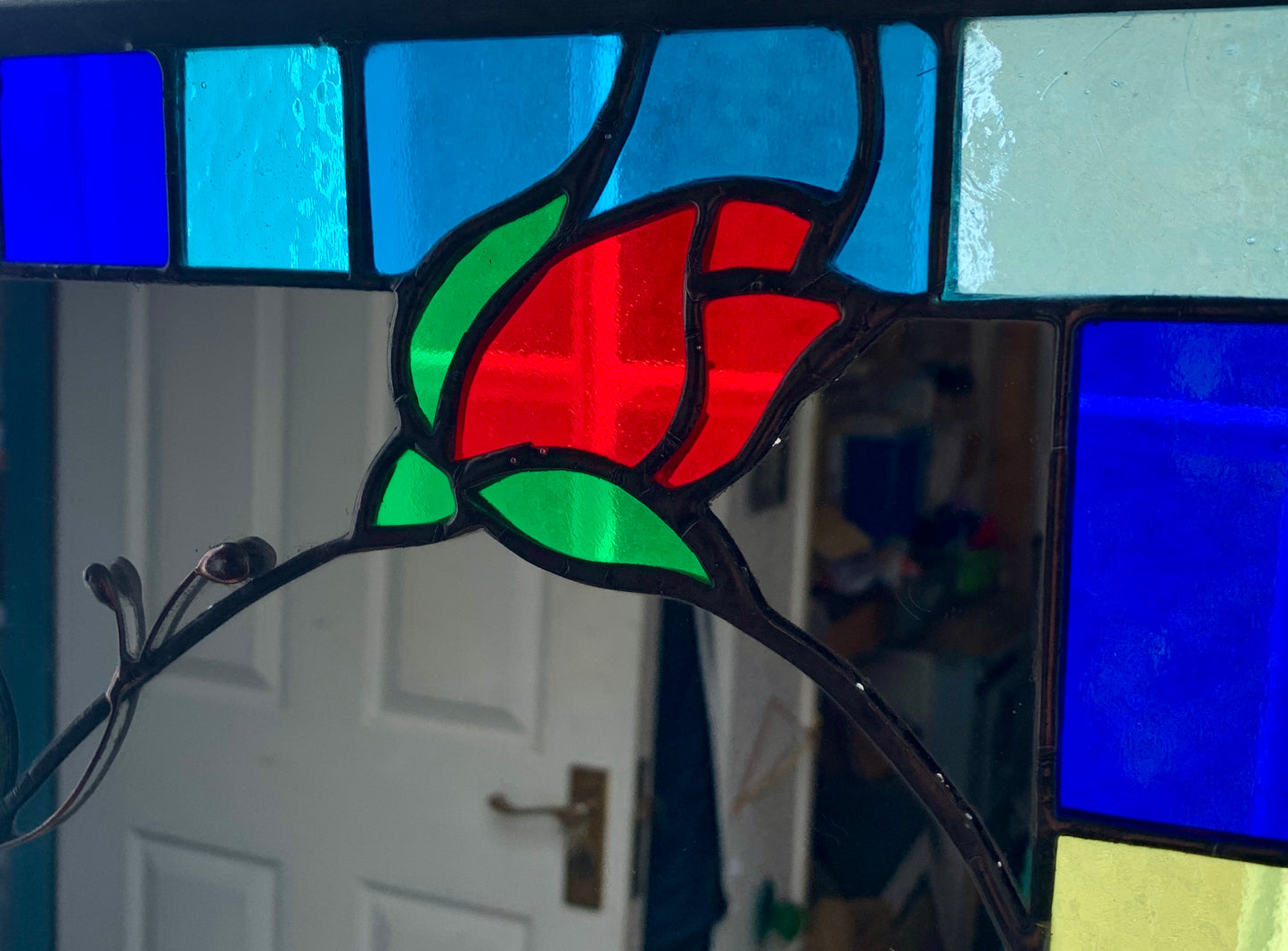 Tall slim stained glass mirror with random colours in various sizes in the border. Red rose bud detail with a long stem cut into the mirror glass and border. Copperwire attached to the back for hanging.