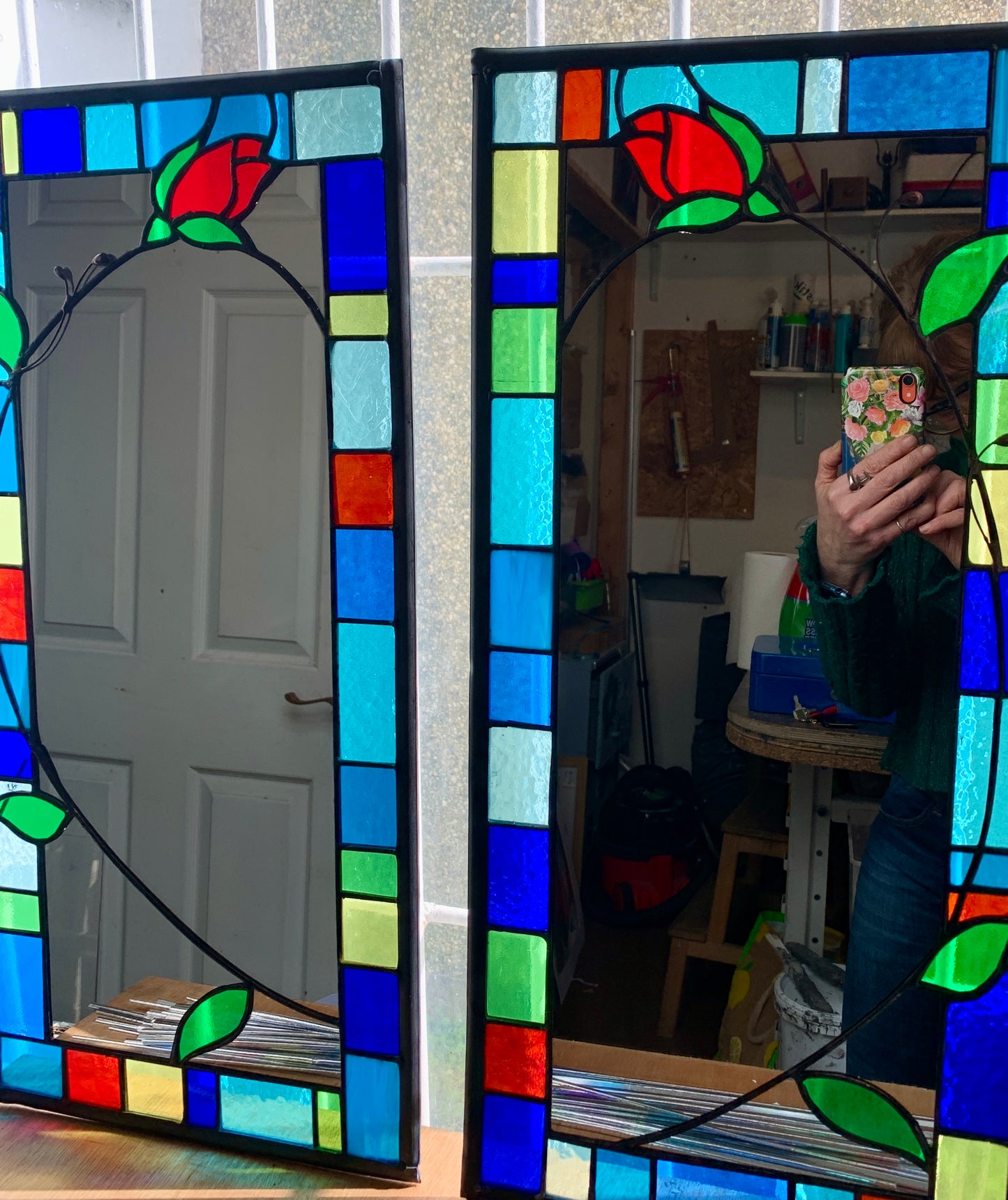 Tall slim stained glass mirror with random colours in various sizes in the border. Red rose bud detail with a long stem cut into the mirror glass and border. Copperwire attached to the back for hanging.