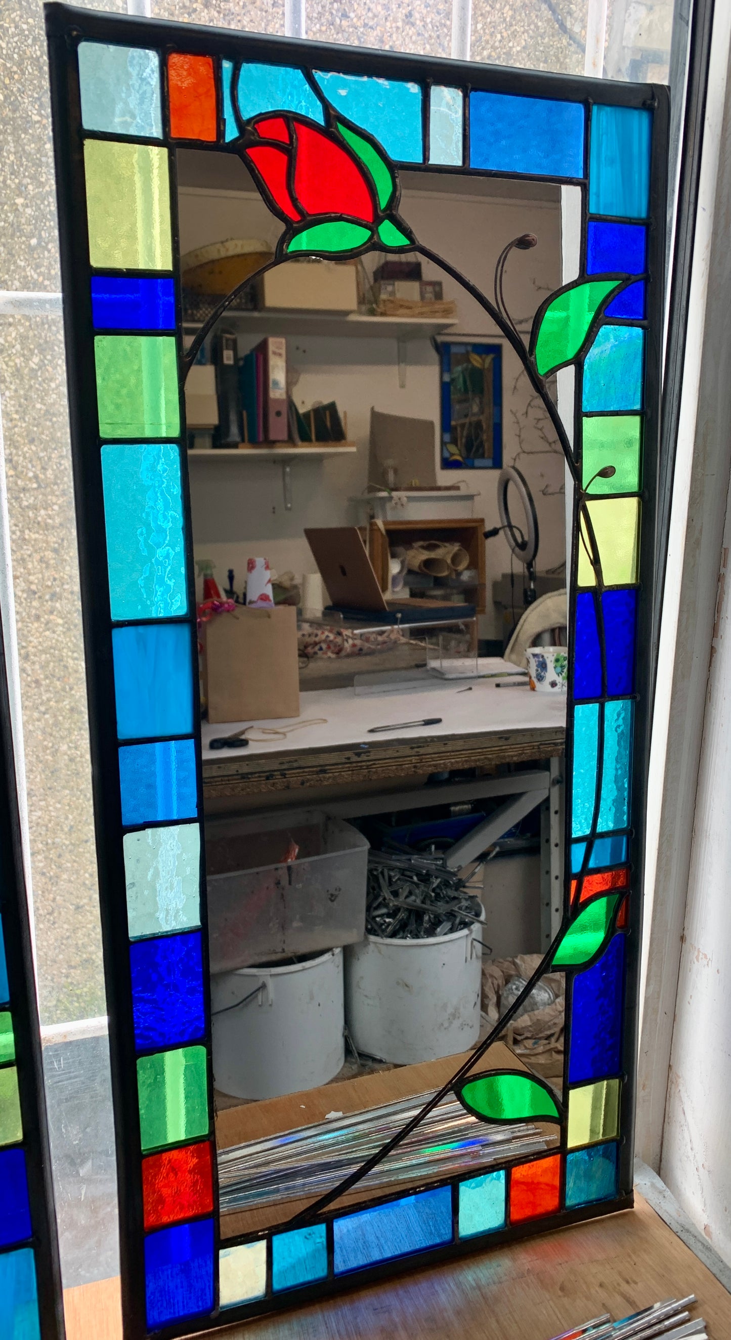 Tall slim stained glass mirror with random colours in various sizes in the border. Red rose bud detail with a long stem cut into the mirror glass and border. Copperwire attached to the back for hanging.