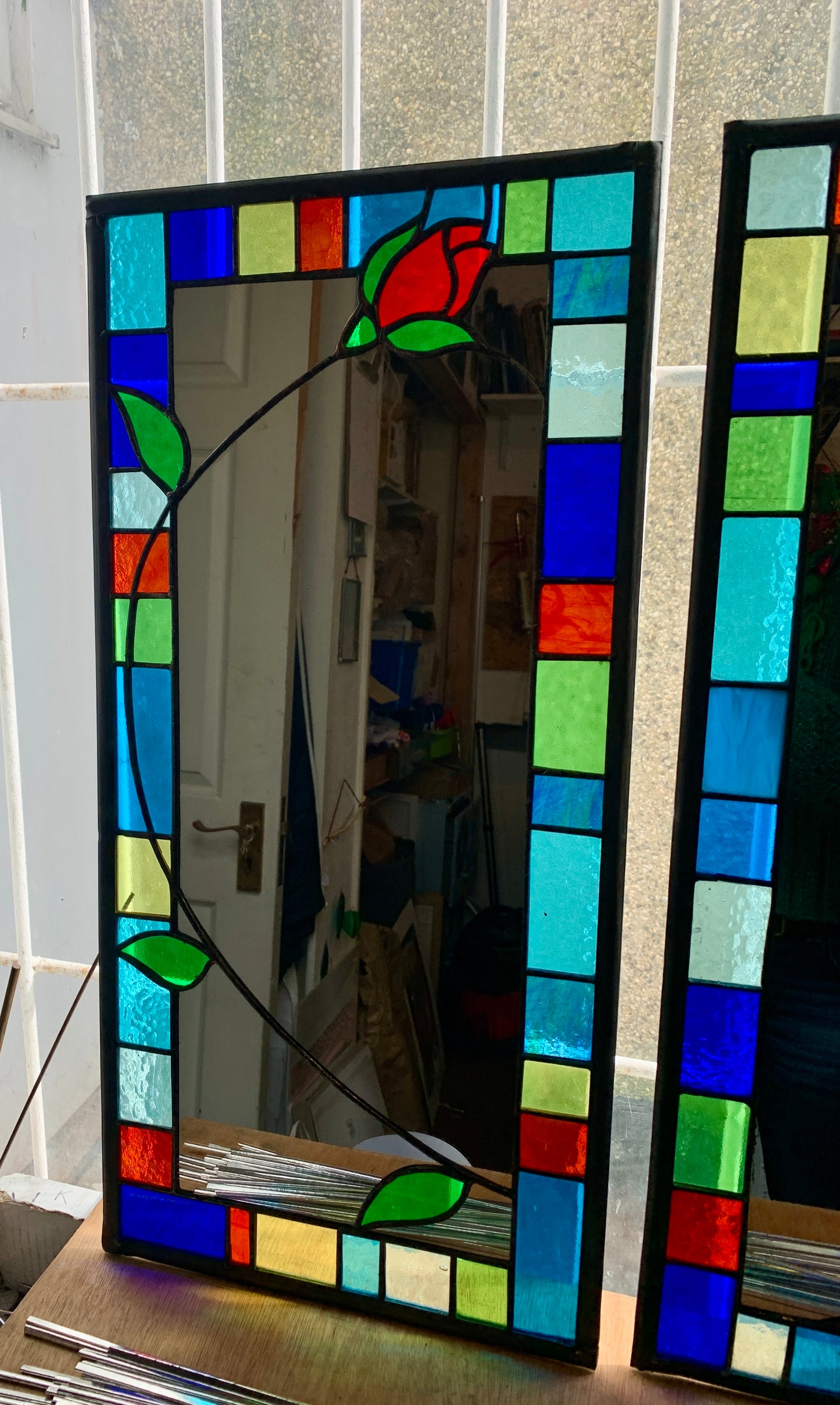 Tall slim stained glass mirror with random colours in various sizes in the border. Red rose bud detail with a long stem cut into the mirror glass and border. Copperwire attached to the back for hanging.
