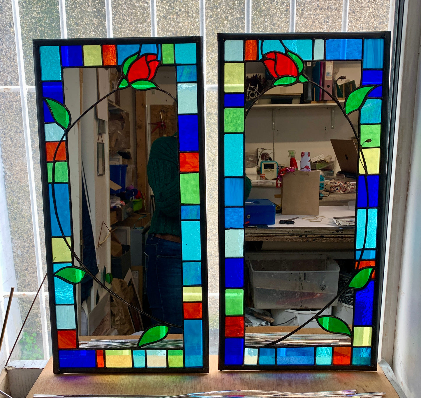 Tall slim stained glass mirror with random colours in various sizes in the border. Red rose bud detail with a long stem cut into the mirror glass and border. Copperwire attached to the back for hanging.