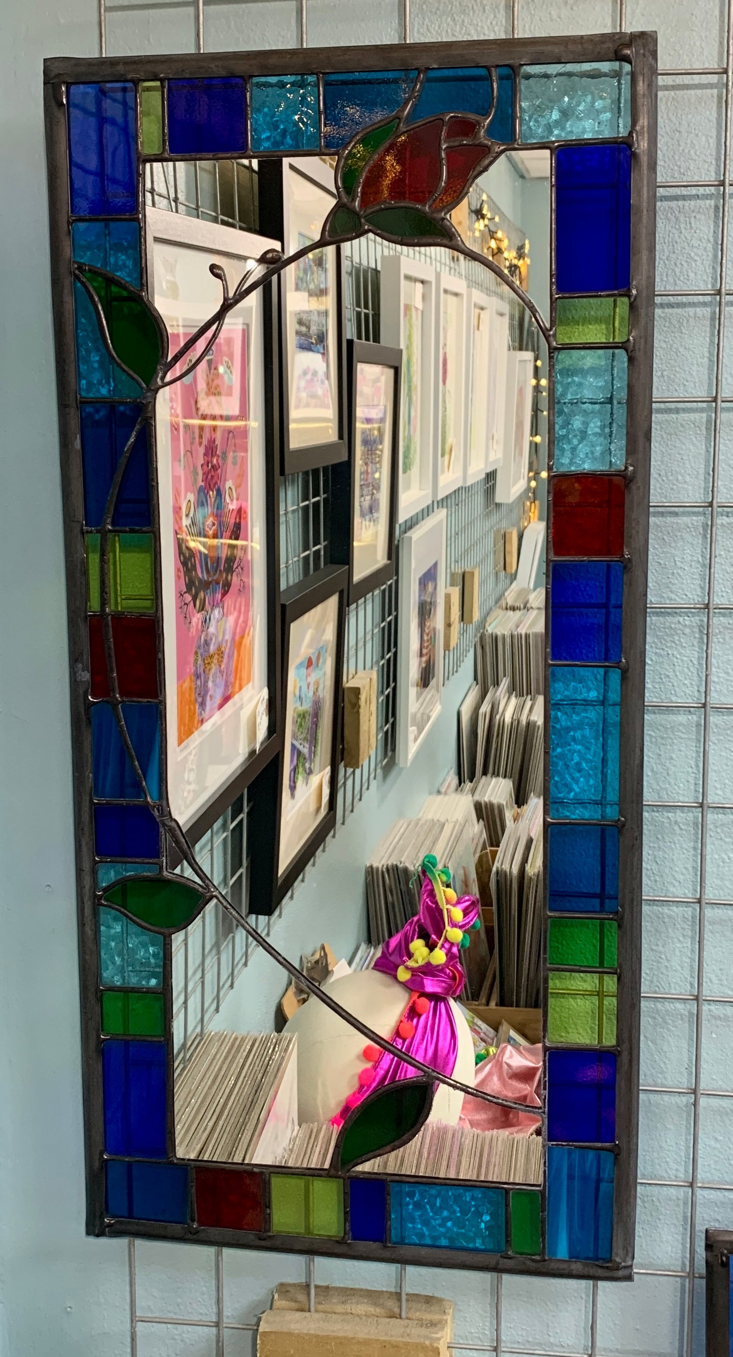 Tall slim stained glass mirror with random colours in various sizes in the border. Red rose bud detail with a long stem cut into the mirror glass and border. Copperwire attached to the back for hanging.