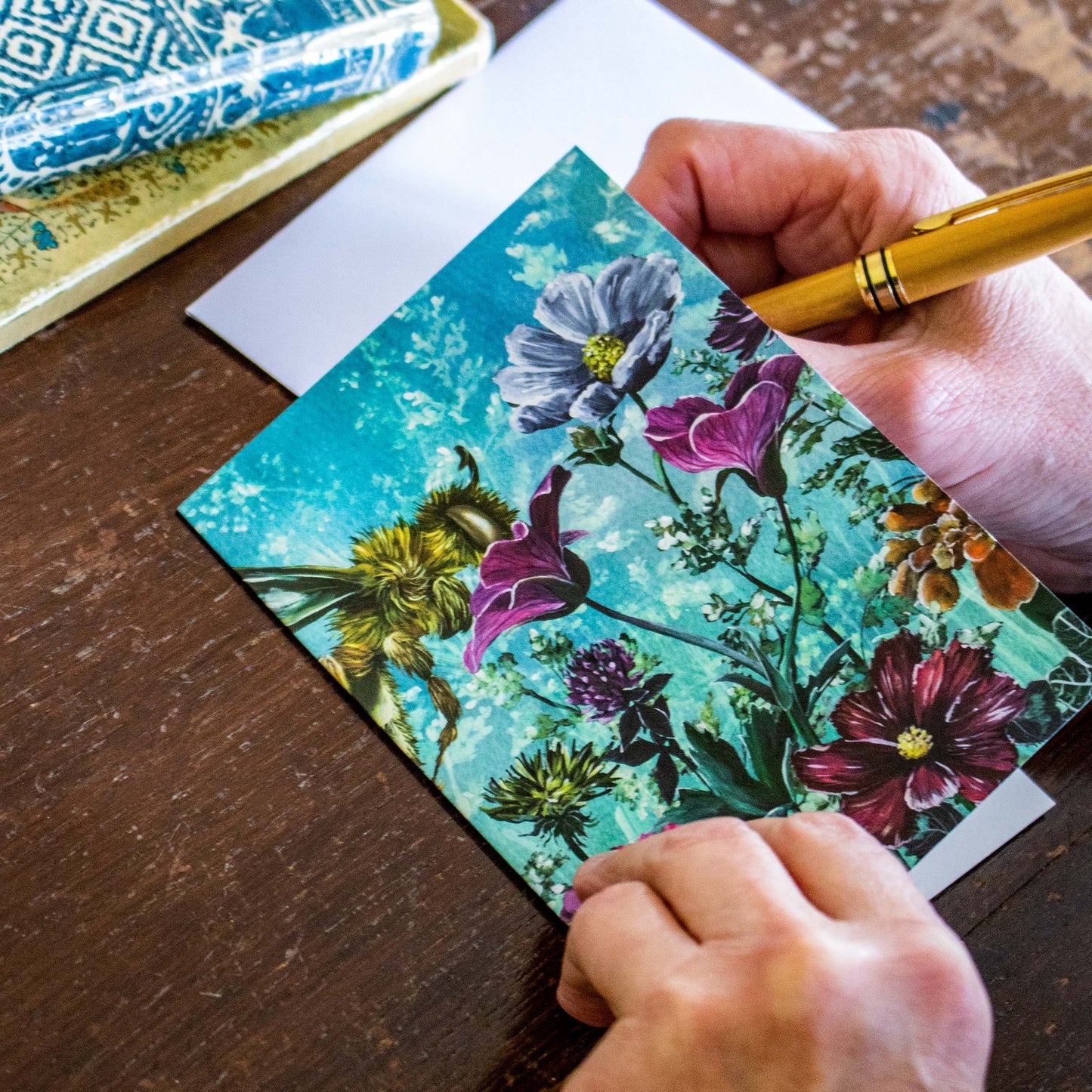 This beautiful floral greeting card features a vast meadow of hand-painted floral acrylic illustrations growing from one side to the other. A wild bee is thoroughly enjoying flying from the pink zinnia, to the red cosmos and purple corn flower that are blooming against a light blue watercolour background.