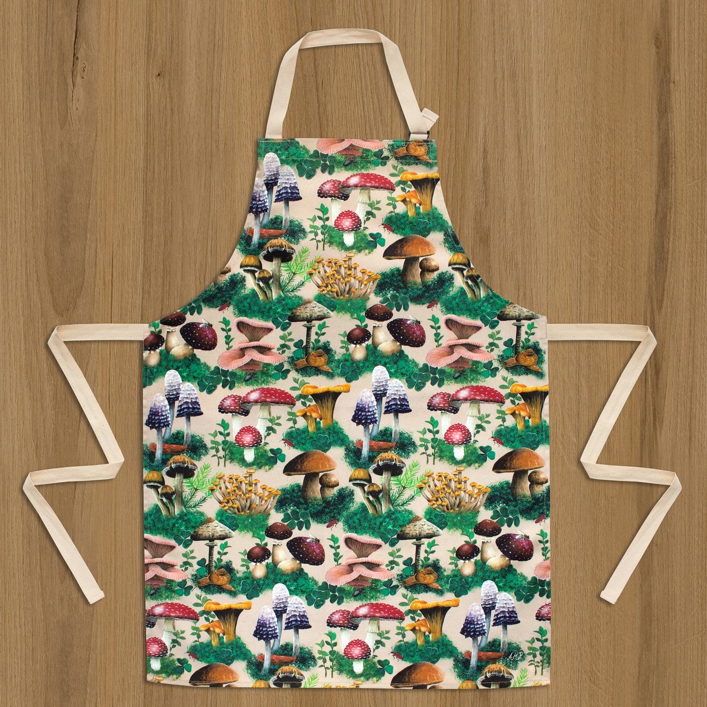 An apron laying flat against a wooden background. The apron's design depicts a variety of woodland mushrooms and foliage against a cream background.