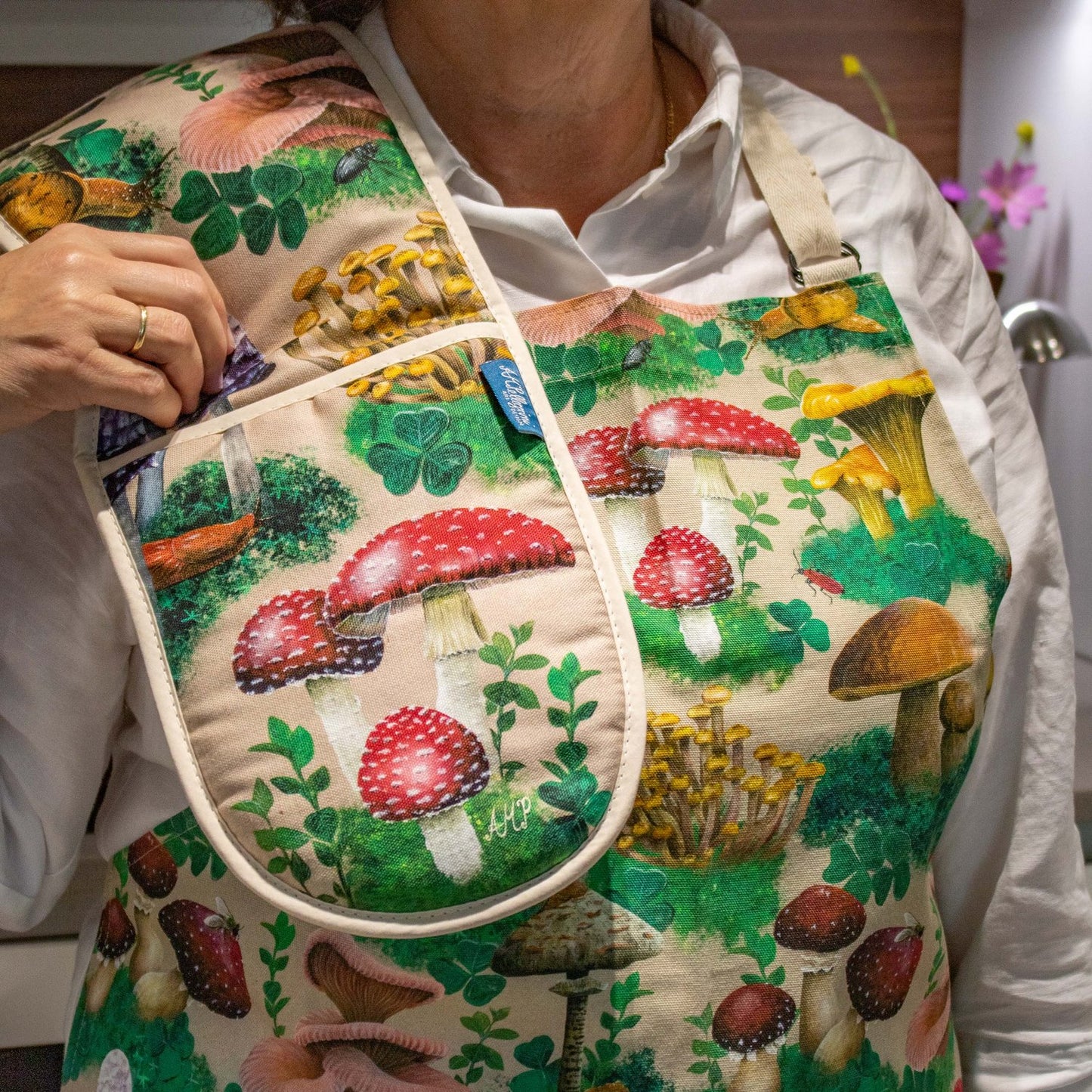 A close up photo of a person's torso wearing an apron with a pair of oven gloves draped over their shoulder. The oven gloves and apron have a design depicting hand-painted woodland mushrooms against a sandy beige background.