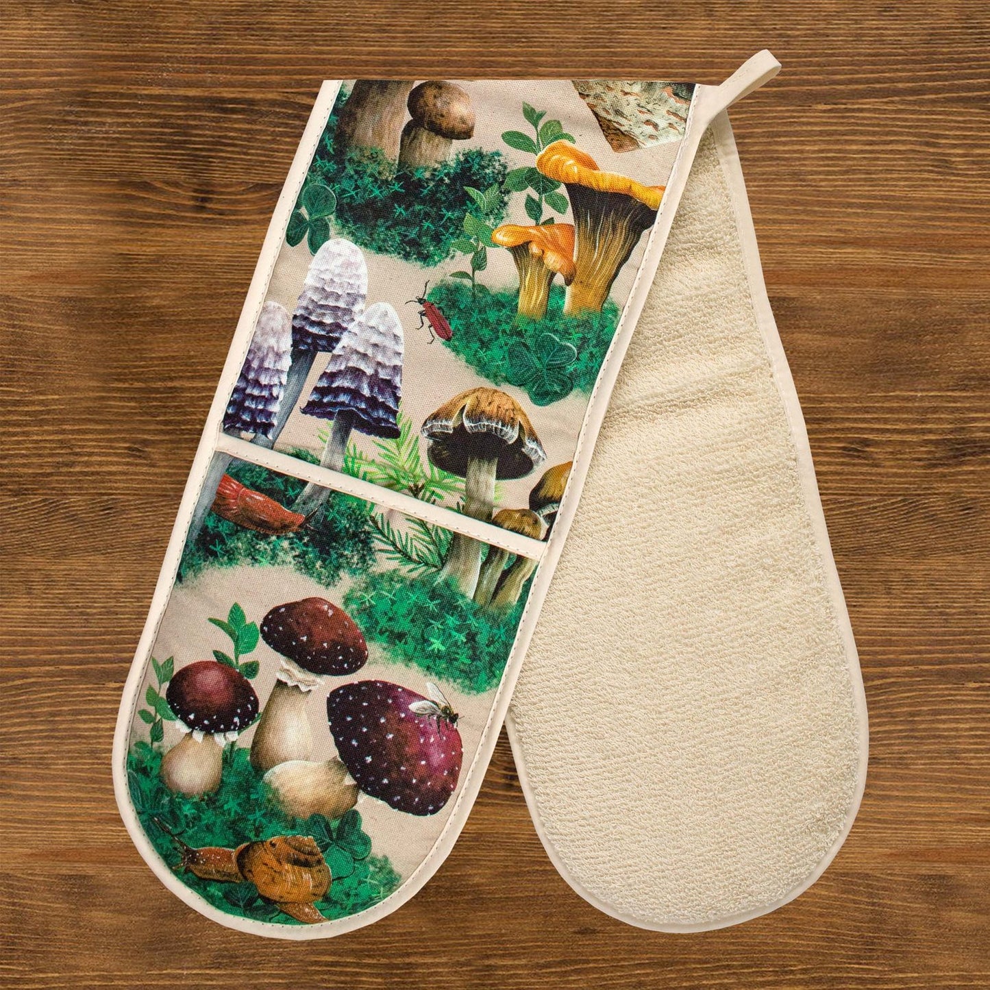 A pair of oven gloves against a wooden background. The oven gloves have a design depicting hand-painted woodland mushrooms against a sandy beige background.