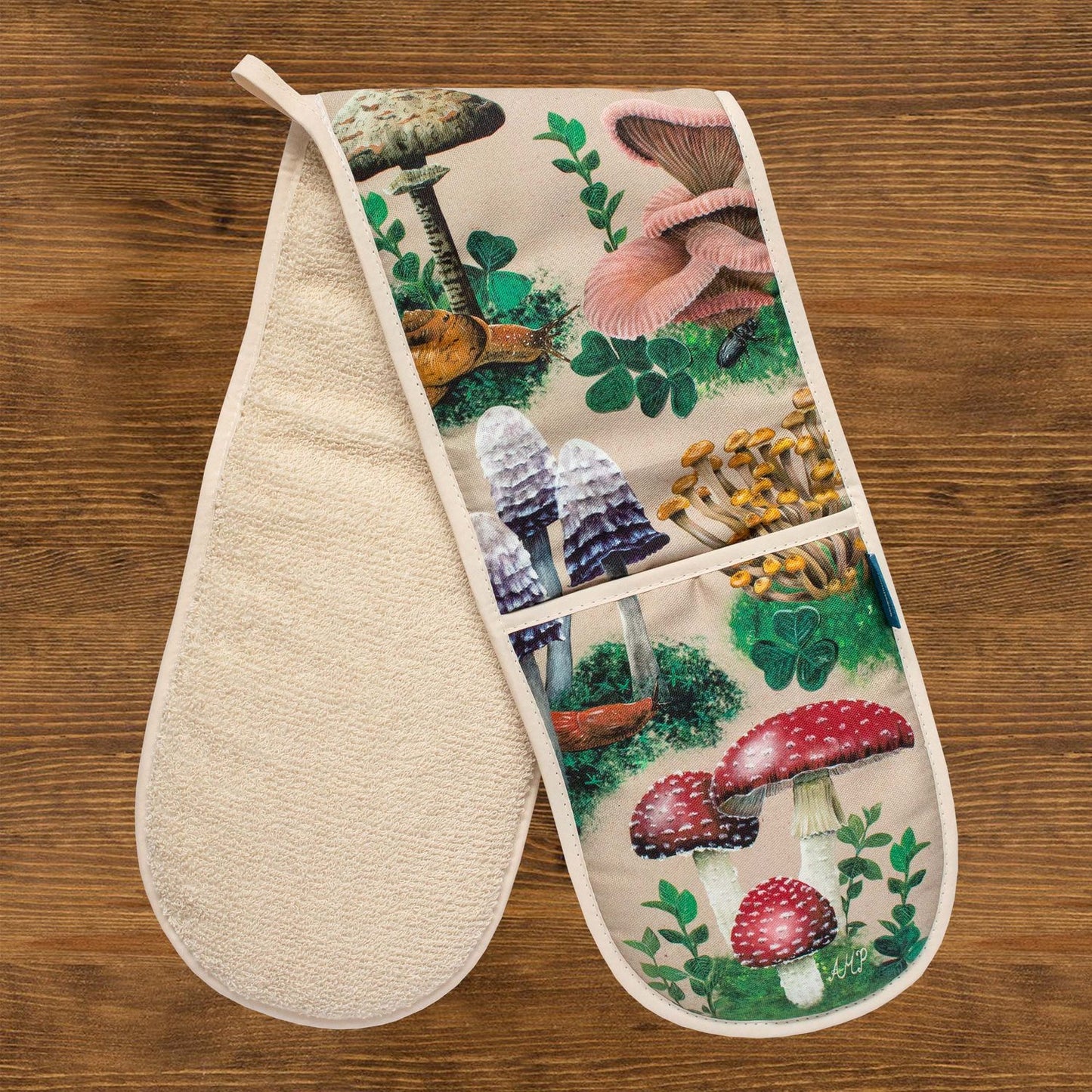 A pair of oven gloves against a wooden background. The oven gloves have a design depicting hand-painted woodland mushrooms against a sandy beige background.