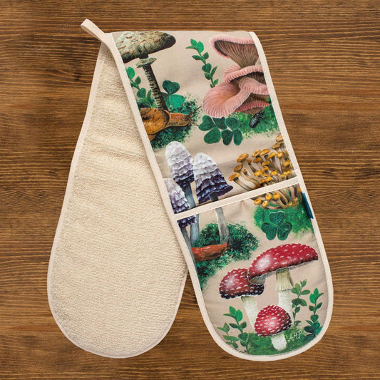 A pair of oven gloves against a wooden background.  The oven gloves have a design depicting hand-painted woodland mushrooms against a sandy beige background.