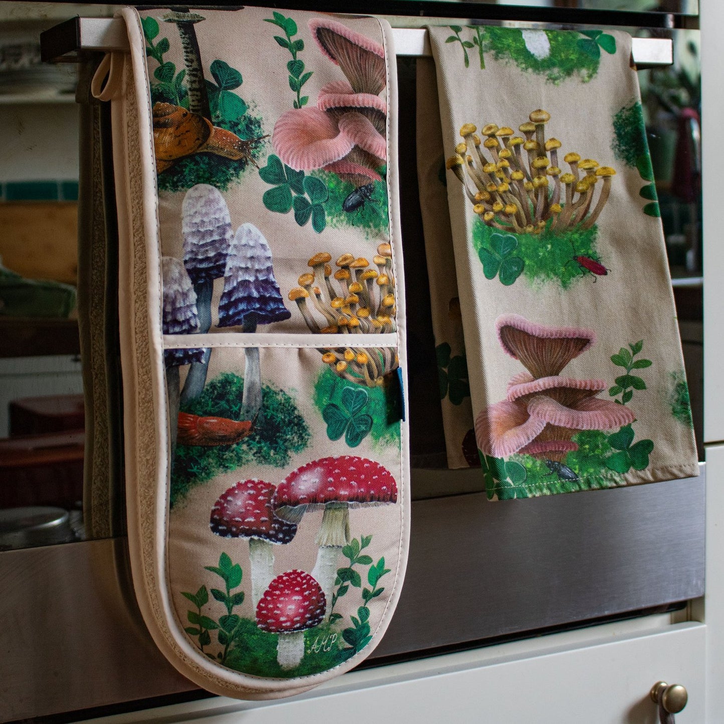 A pair of oven gloves and a tea towel folded over the handle of an oven door. The tea towel and oven gloves have a design depicting hand-painted woodland mushrooms against a sandy beige background.
