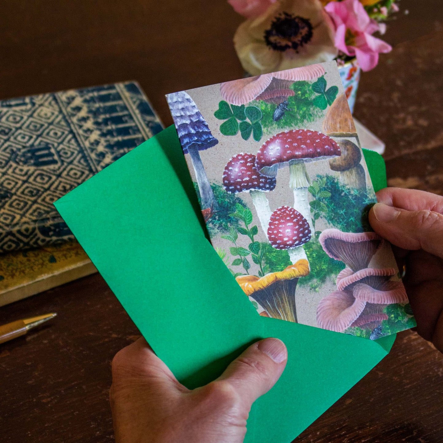 This bold Woodland Shrooms greeting card features a dense design of various mushrooms and fungi, surrounded by patches of moss. It has been printed on a sandy beige background. A bright red toadstool (or Fly Agaric) is sitting right in the middle of the card. The red Fly Agaric is surrounded by Chanterelles, Pink Oyster mushrooms, Inkcap mushrooms and an occasional snail or slug crawling through here and there.
