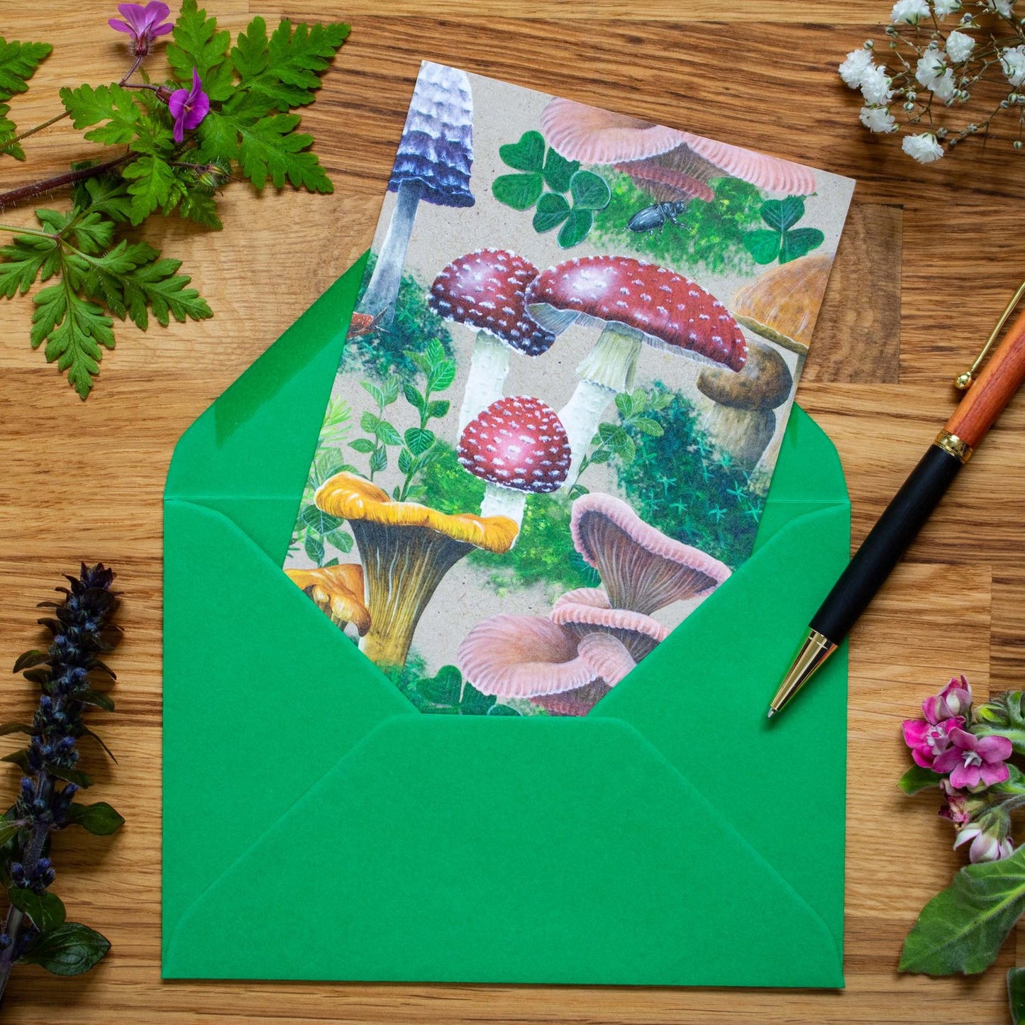 This bold Woodland Shrooms greeting card features a dense design of various mushrooms and fungi, surrounded by patches of moss. It has been printed on a sandy beige background. A bright red toadstool (or Fly Agaric) is sitting right in the middle of the card. The red Fly Agaric is surrounded by Chanterelles, Pink Oyster mushrooms, Inkcap mushrooms and an occasional snail or slug crawling through here and there.