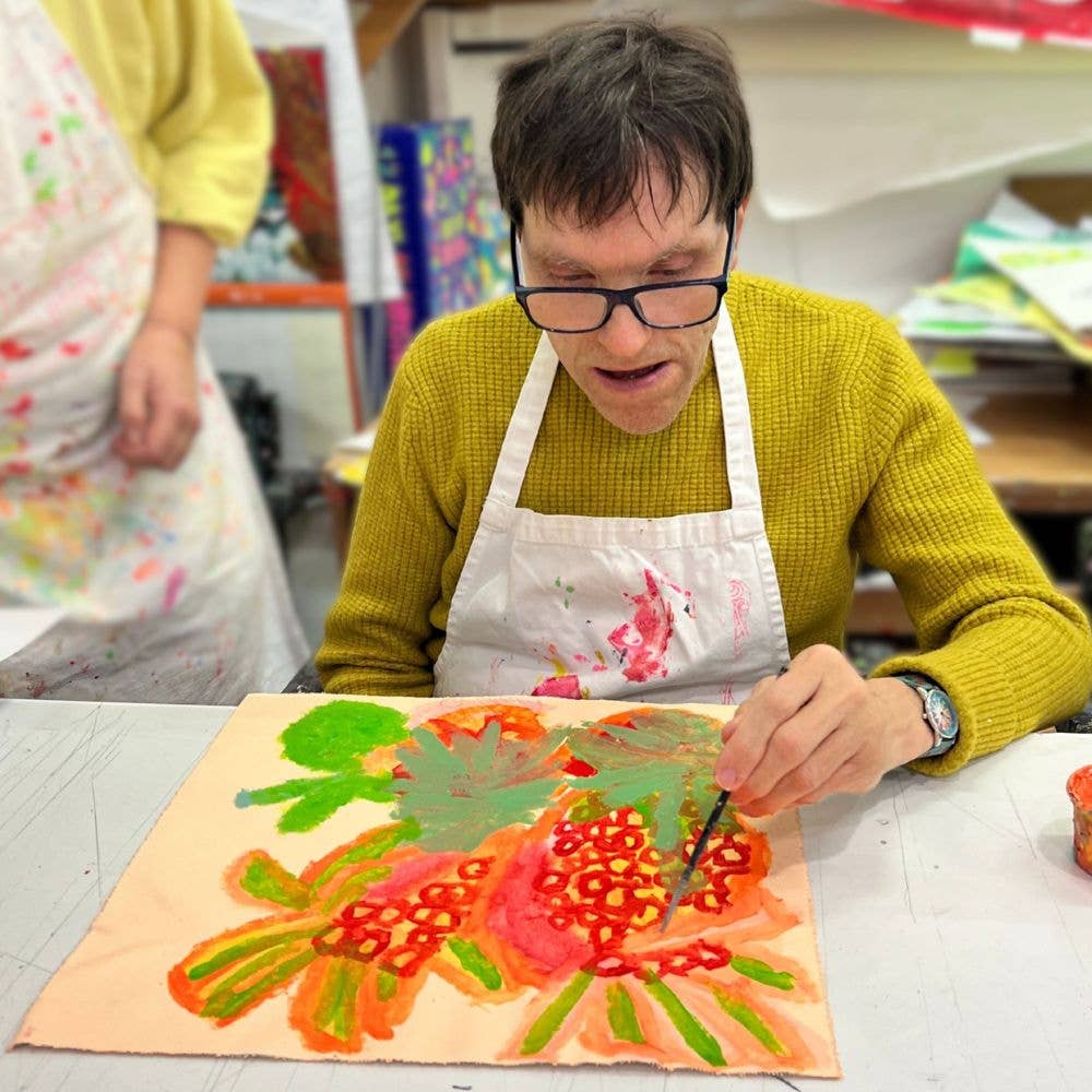 Person painting a colorful pineapple on a piece of paper with another person in the background.