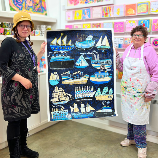 Two people holding a large piece of artwork with boat illustrations in a store setting.