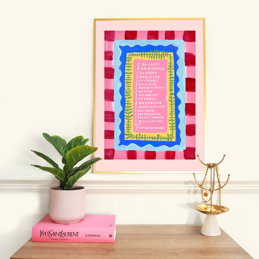 Framed artwork with colorful design on a wall above a shelf with books and a plant.