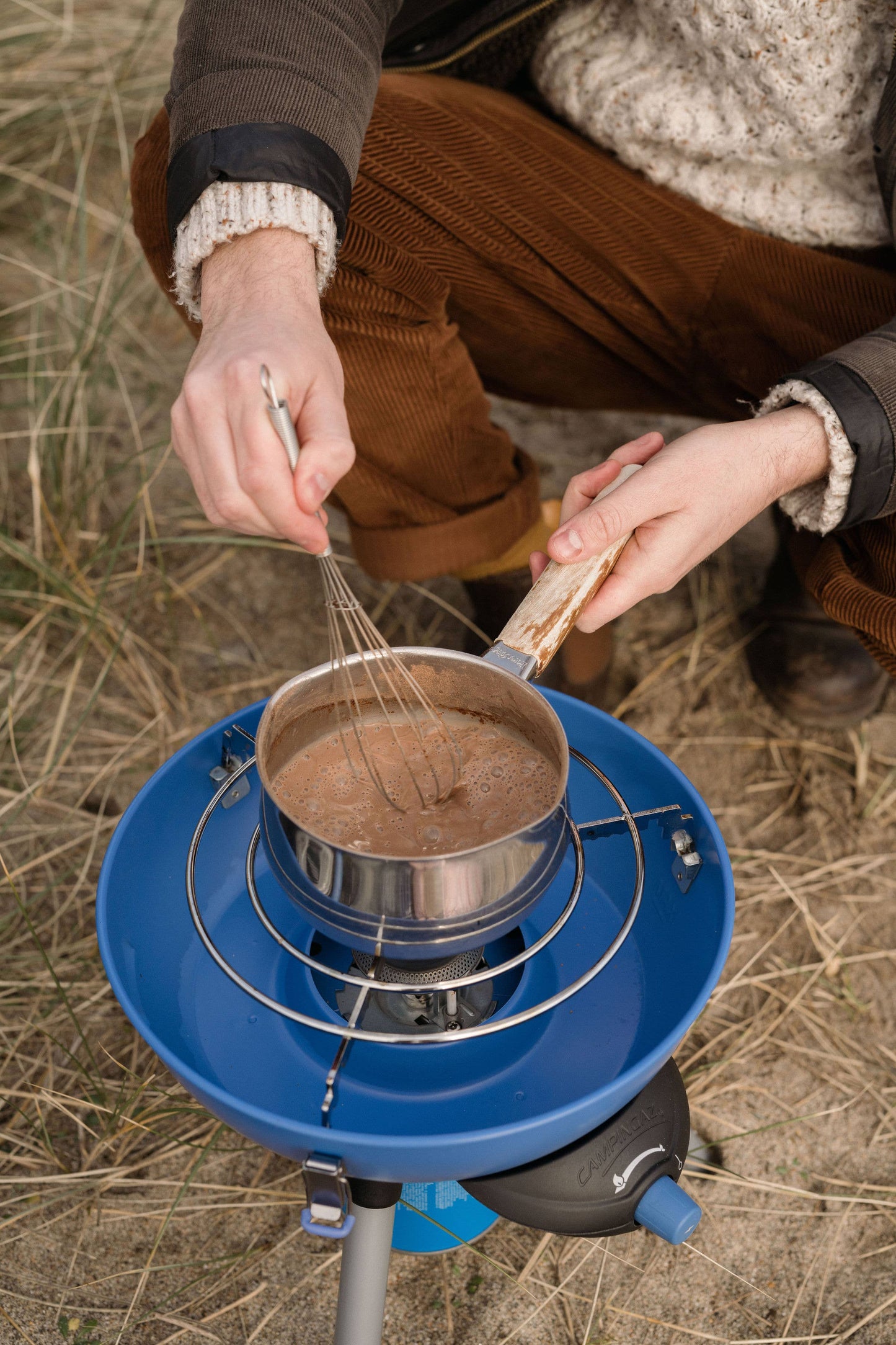 Person cooking outdoors using a portable stove with a pot and whisk