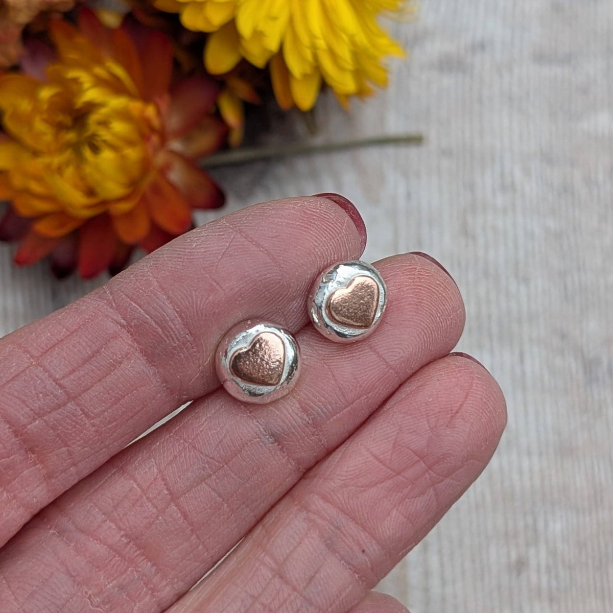 A close-up of a person's hand holding a pair of small round stud earrings with a polished silver outer surface and a smaller rose-gold heart-shaped center, against a light wood background with blurred orange and yellow flowers.