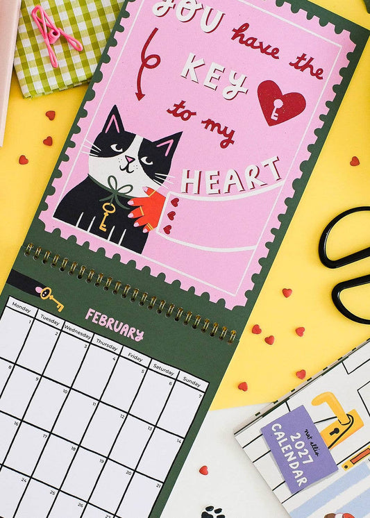 A spiral-bound desk calendar with a pink stamp-like cover featuring a black-and-white cat holding a key and the phrase 'You have the key to my heart.' The calendar is shown open on a yellow surface with scattered red heart cutouts, alongside other stationery items.