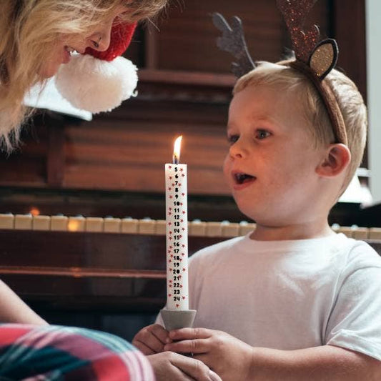 Child wearing reindeer antlers holding a candle with numbers, next to a person in a festive outfit.