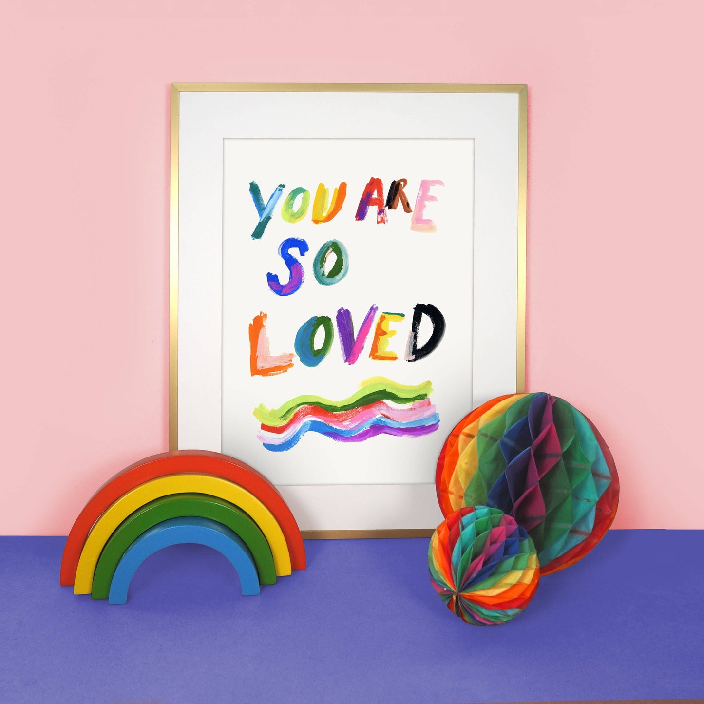 Framed artwork with 'You Are So Loved' text, rainbow decor elements on a pink and blue background