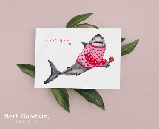 Card with a shark wearing a pink sweater holding a flower, on a pink background with green leaves.
