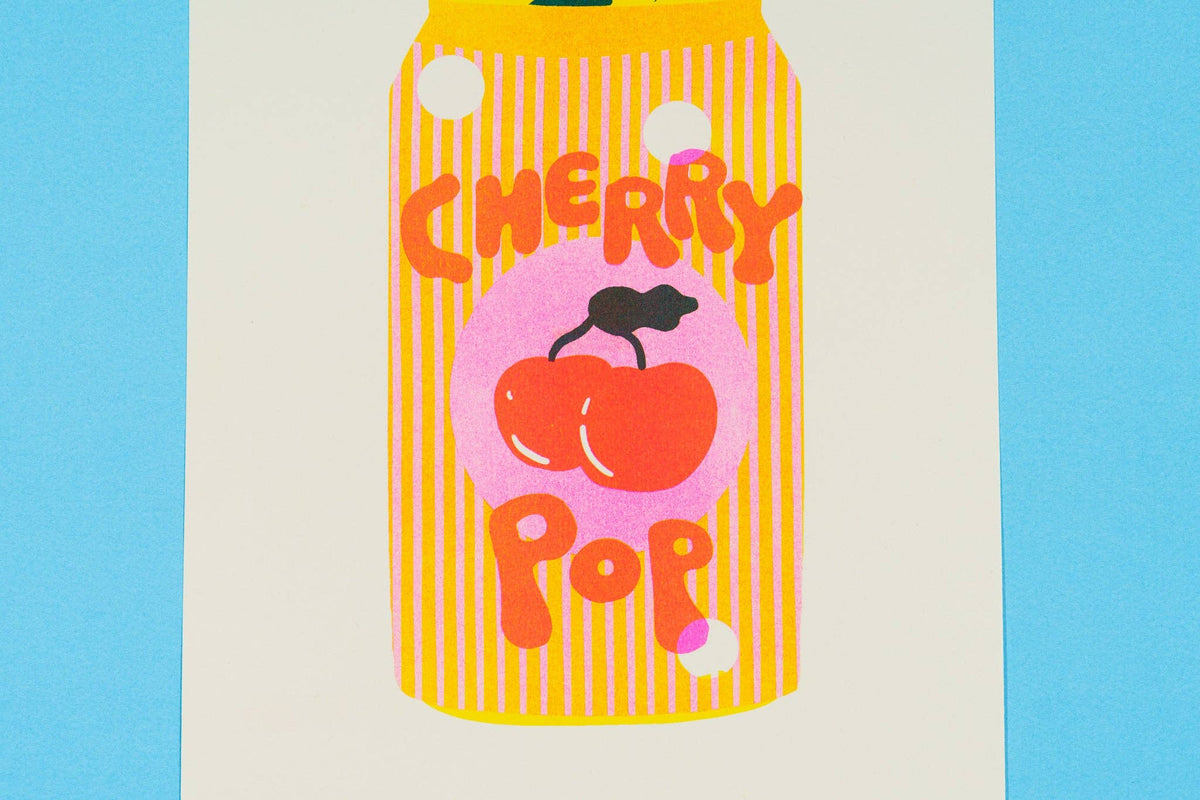 A colorful art print featuring a stylized yellow striped can labeled 'CHERRY POP' with red cherries and a pink circle, displayed on a white sheet against a bright blue background.
