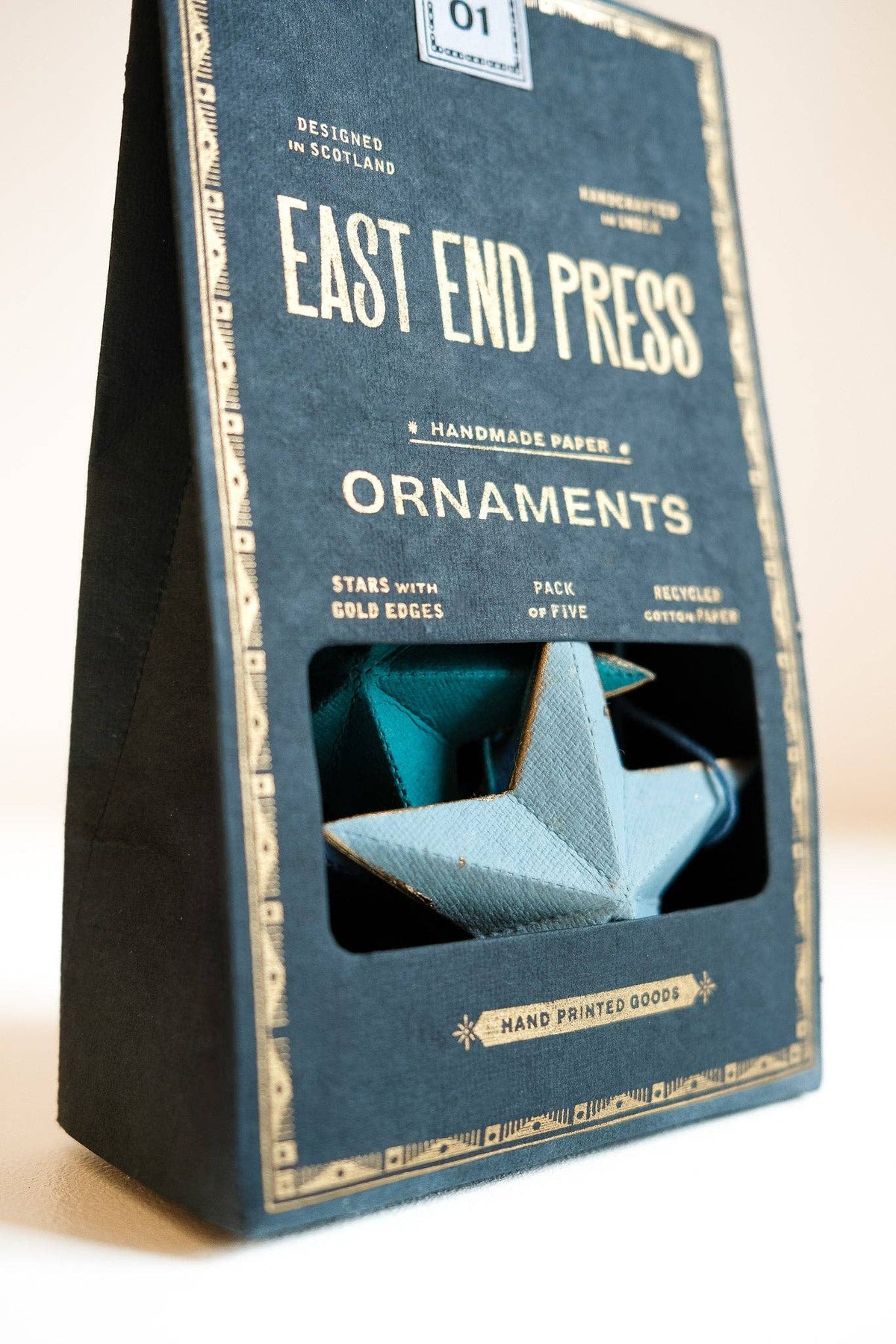 A dark blue paper bag package labeled 'EAST END PRESS ORNAMENTS' with a window showing a folded paper star ornament inside, photographed on a light neutral background.
